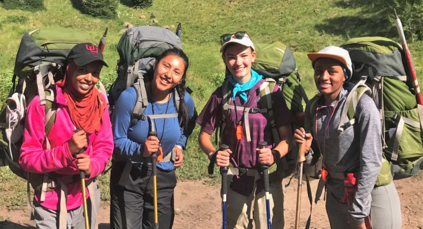 Four people wearing backpacks smile for a photo in a green area